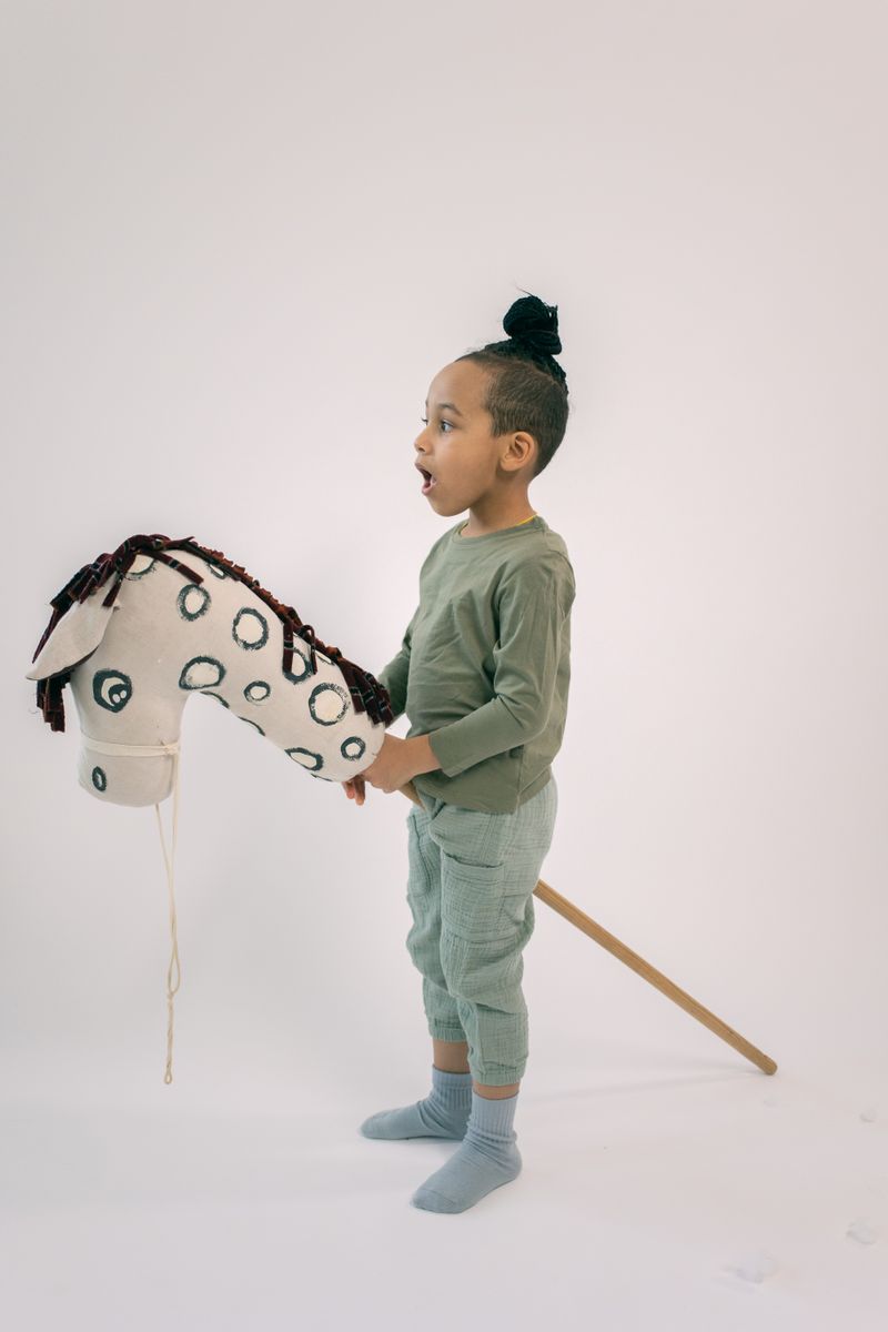Side view of cute boy in stylish outfit with high bun riding stick horse on white background