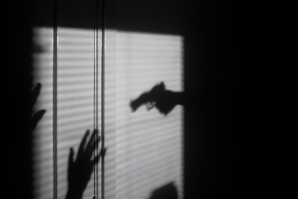 Shadows of hands and a handgun cast against a window with closed blinds