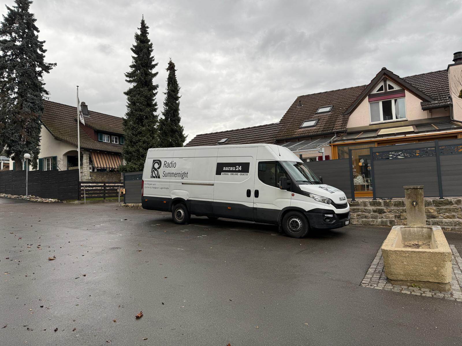 Livesendung Schule Umiken 19.12.2024 | Weißer Transporter von Radio Summenlicht auf einem Parkplatz, umgeben von Bäumen und Wohnhäusern.
