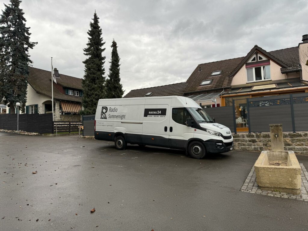 Weißer Transporter von Radio Summenlicht auf einem Parkplatz, umgeben von Bäumen und Wohnhäusern.