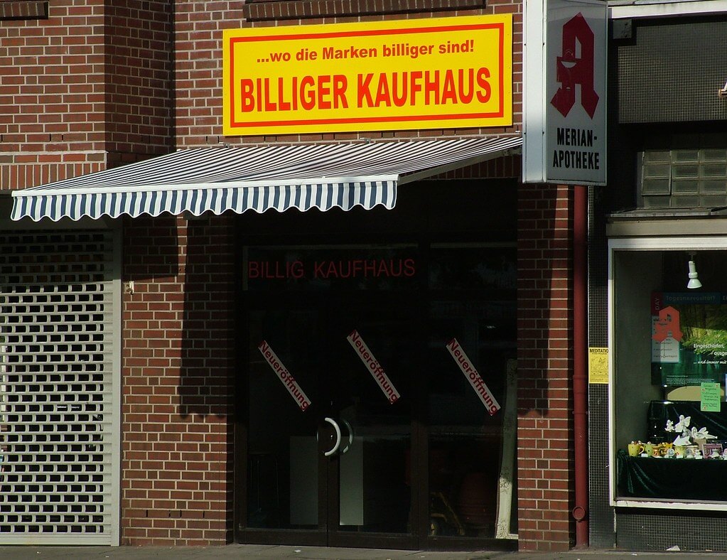 Eingang eines Billigen Kaufhauses mit gelbem Schild und Markisen, das Rabatte bewirbt.