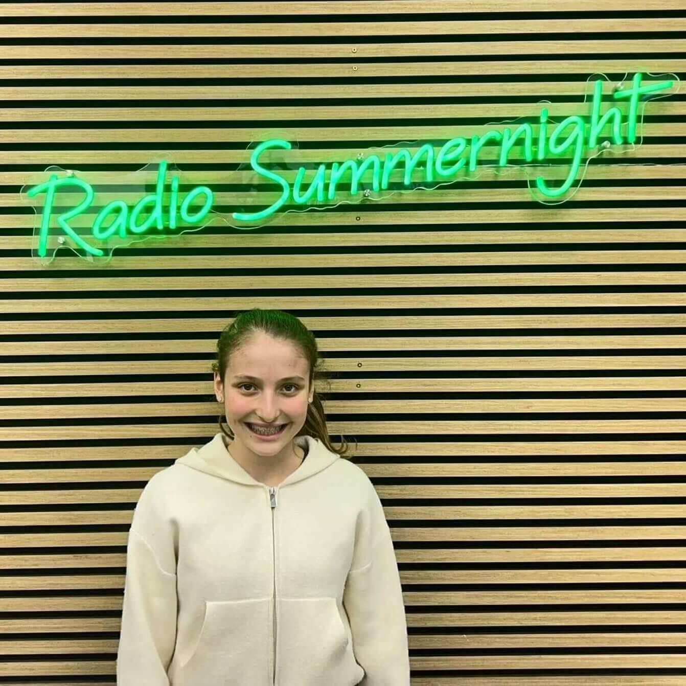 Ein Mädchen steht lächelnd vor einem Neon-Schild mit der Aufschrift Radio Summernight, das in grün leuchtet. Holzlatten im Hintergrund.